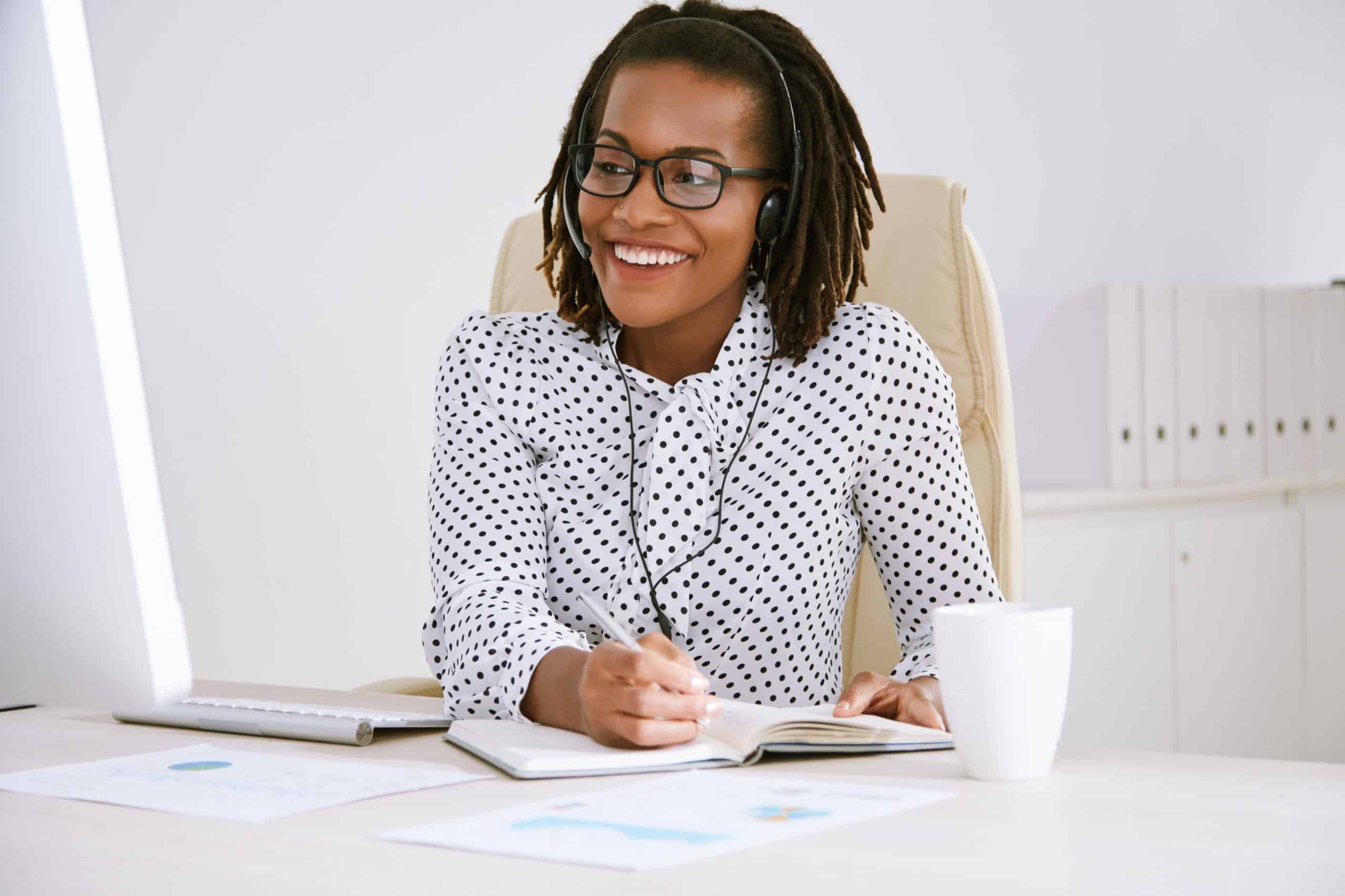 Cheerful smiling female entrepreneur using headset to talk to coworker and taking notes